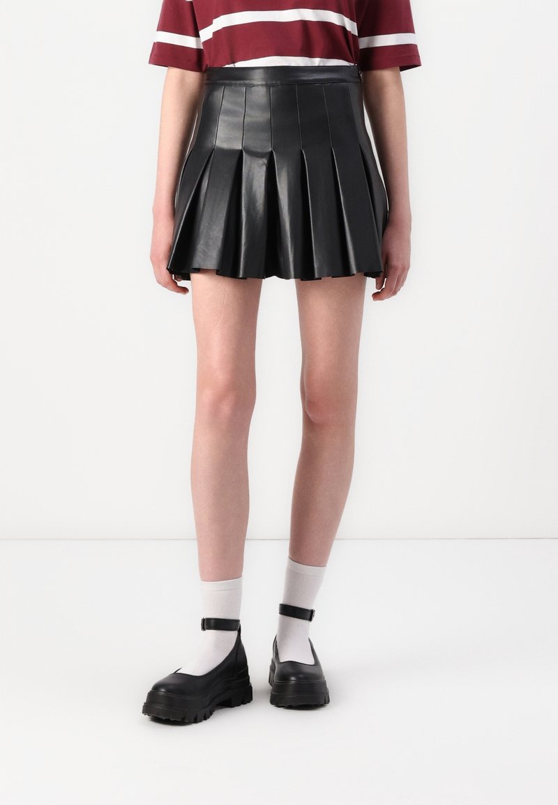 Personne portant une mini-jupe en cuir noire plissée, des chaussettes blanches aux chevilles, des chaussures Mary Jane noires et une chemise rouge à rayures blanches.