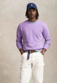 Sweatshirt lavanda com punhos canelados, calças brancas e um cinto multicolorido. Os acessórios incluem um boné azul e um relógio.