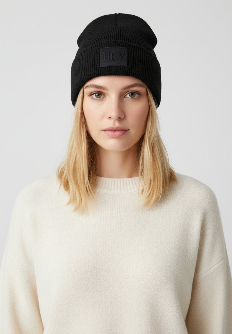 Zwarte gebreide beanie met een geribbelde textuur en een rechthoekige zwarte patch waarop "HEY" staat, gedragen met een crèmekleurige trui.