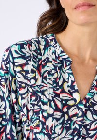Blusa floreale blu navy con vivaci motivi bianchi, rossi e verdi, un colletto con bottoni e un tessuto morbido e texturizzato con dettagli plissettati.