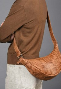 Bolso de hombro de cuero marrón con forma curvada, superficie texturizada y correa ajustable. Cerrado con un detalle de hardware metálico.