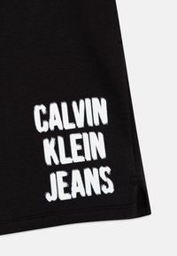 Czarne szorty materiałowe z białym logo „CALVIN KLEIN JEANS” w pogrubionych, blokowych literach na dolnej lewej stronie.