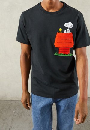 Person trägt ein schwarzes T-Shirt mit einer Grafik von Snoopy und Woodstock auf einer roten Hundehütte-Tasche, kombiniert mit blauen Jeans.