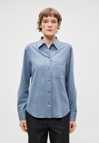Svetle modrá košile na knoflíky z měkké látky s límcem, dlouhými rukávy, přední kapsou a mírně zaobleným spodním okrajem.