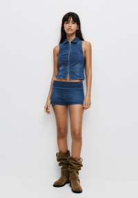 Denim crop top with collar and front zipper, paired with matching denim shorts. Model wears slouchy brown suede boots. White background.