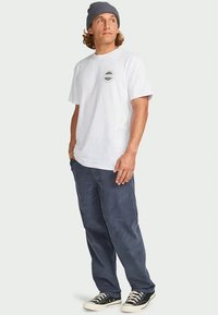 White cotton t-shirt with a circular graphic on the chest, paired with navy corduroy pants and black sneakers. Gray beanie on head.