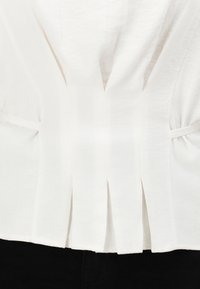 Top bianco in lino con un design plissettato, legato ai lati e con scollatura a V, che offre un aspetto leggero e di consistenza.