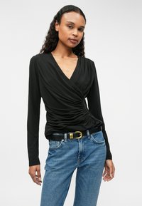 Melnā garo piedurknu apģērba novilkta augšdaļa ar drapētu auduma dizainu, ar V veida izgriezumu un sakārtotu akcentu sānos, apvienota ar augsta vidukļa ziliem džinsiem.