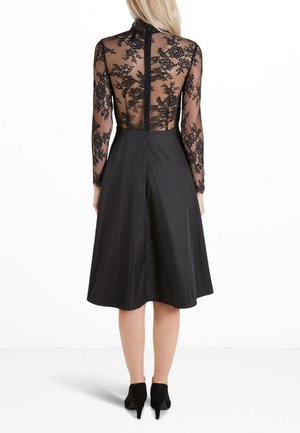 Schwarzes Kleid mit einem figurbetonten Spitzenoberteil mit langen Ärmeln und einem ausgestellten Rock. Verschluss hinten mit Reißverschluss, mittellang bis zur Wade und mit geschichtetem Stoff.