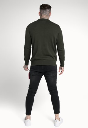 Mężczyzna z tatuażami na szyi i rękach, ubrany w ciemnozielony sweter, czarne obcisłe dżinsy i białe trampki, stojący tyłem na jednolitym tle.
