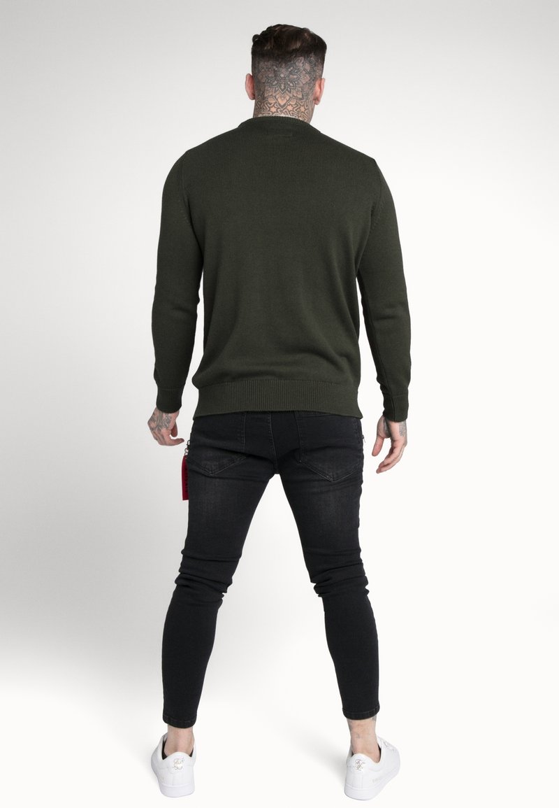Homme avec des tatouages au cou et sur la main, portant un pull vert foncé, un jean skinny noir et des baskets blanches, debout de dos sur un fond uni.