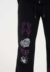 Zwarte sweatpants met paarse tribale ontwerpen en een zilveren schedelafbeelding versierd met rhinestones. Opmerkelijke klauwenafbeelding op het onderbeen.