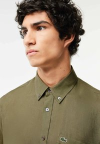 Camicia in cotone verde oliva con bottoni, colletto, chiusura a due bottoni e una piccola etichetta logo sul petto. Tessuto morbido e vestibilità classica.