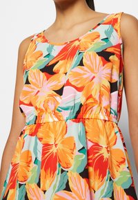 Robe sans manches avec un grand motif floral aux couleurs vives orange, rose, rouge et vert, portée par une personne aux cheveux foncés attachés en arrière.