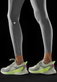 Szare legginsy mają teksturowany wzór oraz białe akcenty w postaci kropek wzdłuż dolnej części nogawek. Jasnoszare buty sportowe w kolorze neonowej zieleni z nakrapianymi podeszwami.