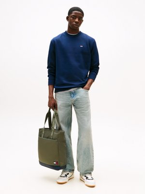 Jonge man in marineblauwe sweatshirt, lichtblauwe spijkerbroek, witte sneakers die een olijfgroene tote bag met merkstraps vasthoudt.
