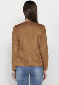 Femme aux longs cheveux bruns portant une veste en daim marron et un jean bleu, vue de dos sur un fond blanc uni.