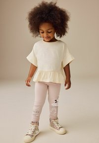 Top peplum color crema chiaro con maniche corte, abbinato a leggings rosa pallido con un design a tema circo e sneakers alte con glitter argento.