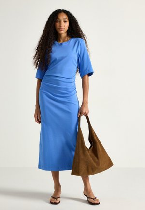 Femme aux cheveux bouclés porte une robe midi bleue et des sandales noires, tenant un grand sac en daim marron devant un fond uni.