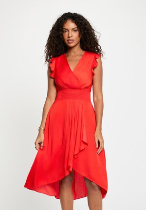 Femme aux cheveux bouclés portant une robe rouge portefeuille sans manches avec des manches à volants et un ourlet asymétrique, debout sur un fond blanc.