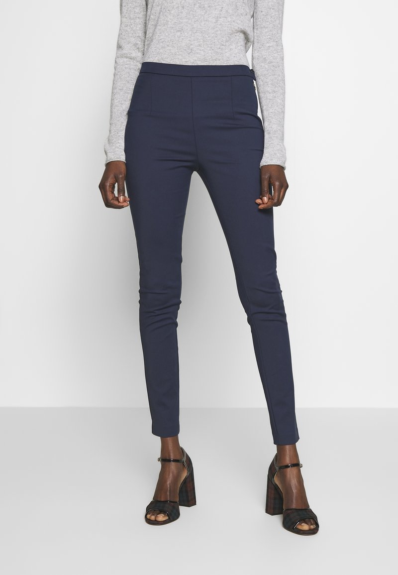 Pantalon bleu marine ajusté avec une taille haute, deux poches avant et une texture lisse, associé à des talons ouverts noirs à carreaux.