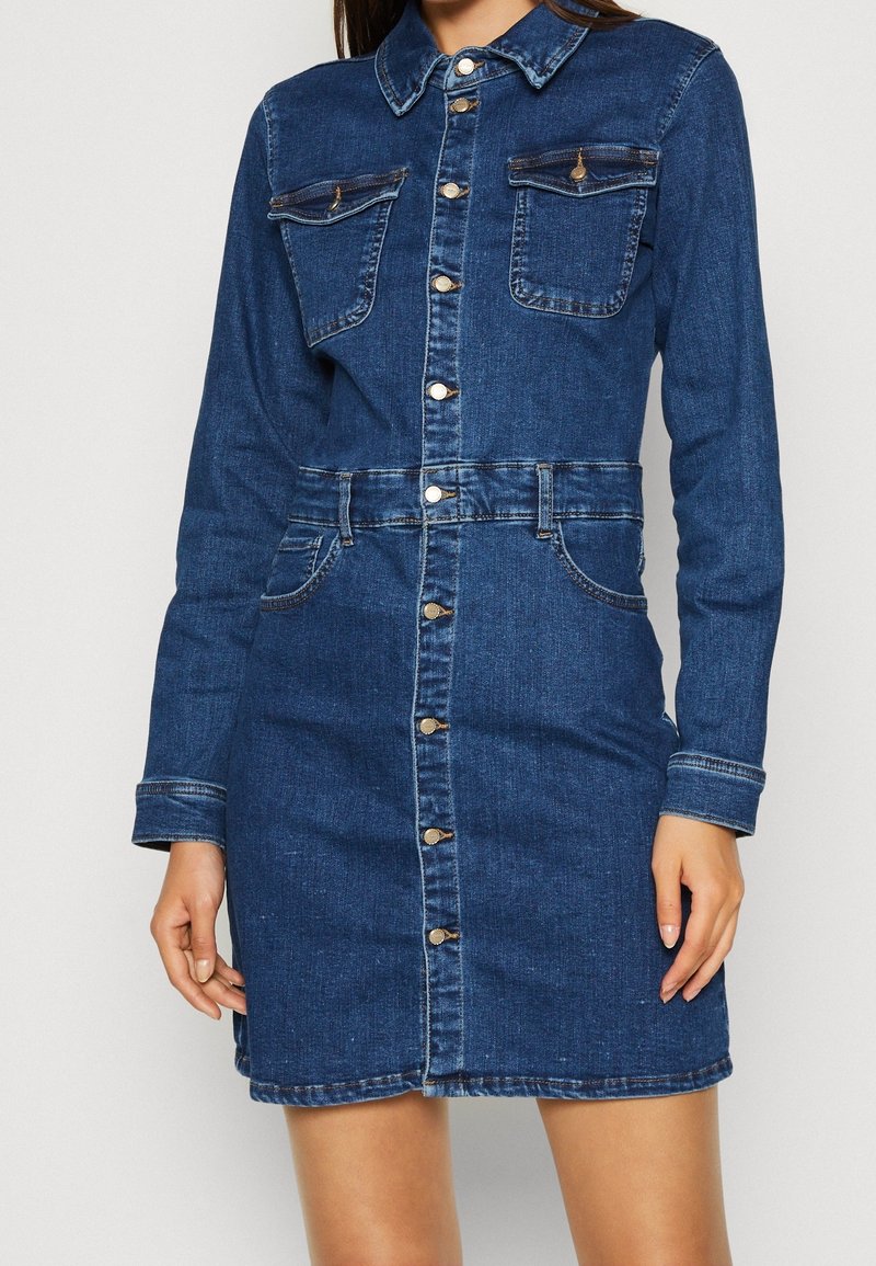Femme portant une robe en jean à manches longues avec boutons, poches à rabats sur la poitrine et détails de couture à la taille à l'avant.