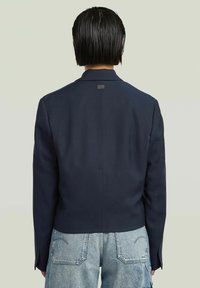 Blazer azul marino con un diseño elegante, ajuste a medida y líneas de hombros estructuradas. Tejido suave y detalle minimalista del logo en la parte trasera.