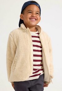 Hellbeige Fleecejacke mit weicher Textur, Reißverschluss vorne und Seitentaschen, getragen über einem weißen gestreiften Hemd mit kastanienbraunen Linien.