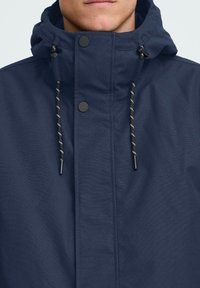 Mornarsko modra jakna z visokim ovratnikom, z gumbi in zadrgo, kapuco na vrvico in kontrastnimi črnimi in belimi črtastimi vrvicami.