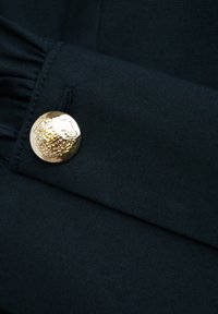 Czarny materiał z zagiętym brzegiem, ozdobiony okrągłym, wypukłym złotym guzikiem o misternych detalach. Tekstura wydaje się gładka i dopasowana.