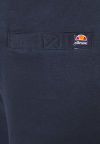 Tissu bleu marine avec détail de boucle rectangulaire cousue et une petite étiquette du logo de la marque Ellesse en blanc, orange et rouge sur le côté droit.