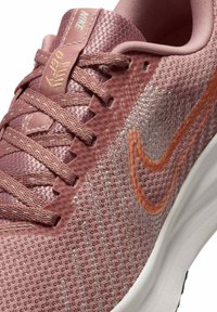 Zapatilla deportiva rosa con una parte superior de malla texturizada, acentos plateados y un prominente logo de swoosh naranja. Presenta cordones planos y una suela acolchada.