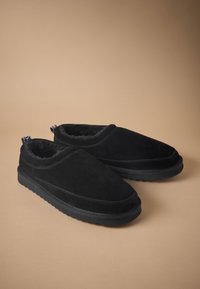 Chaussons en suède noir doux avec une doublure en peluche, profil bas, bout arrondi et semelle en caoutchouc texturée. Présentent des languettes à l'arrière pour faciliter l'enfilage.
