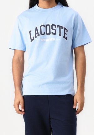 Jasnoniebieska bawełniana koszulka z krótkim rękawem, z wyraźnym granatowym logo z napisem „LACOSTE” oraz umieszczonym pod nim napisem „SPORTSWEAR”.