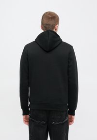 Felpa nera con tessuto strutturato, con cappuccio e polsini a coste, indossata con jeans neri strappati, vista da dietro.