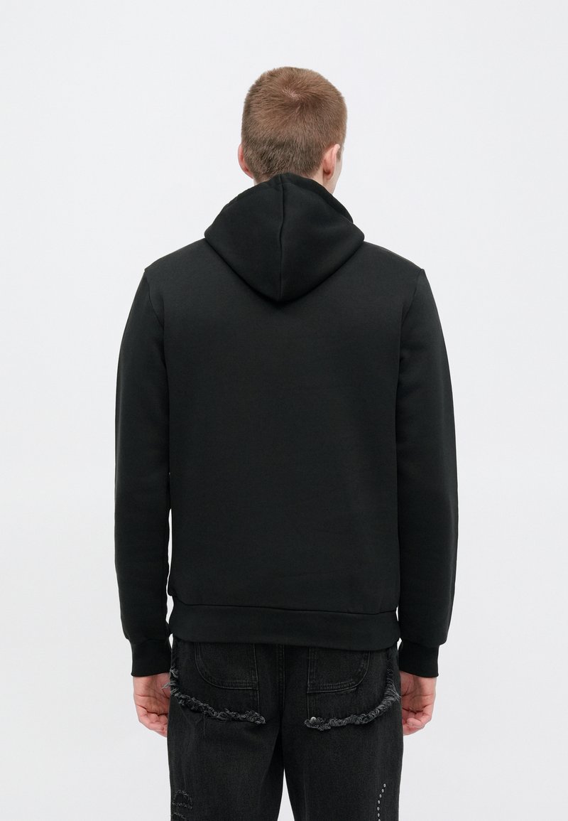 Black hoodie with a textured fabric, featuring a hood and ribbed cuffs, worn with distressed black jeans, seen from the back.