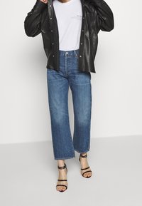 Person i blå vida jeans, svarta klackskor med remmar, vit t-shirt med tryck, och svart läderjacka med tryckknappar.