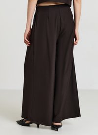 Pantalons à larges jambes en tissu brun foncé, avec des plis et une texture lisse. Portés avec des chaussures à talons noirs, mettant en valeur la coupe élégante.