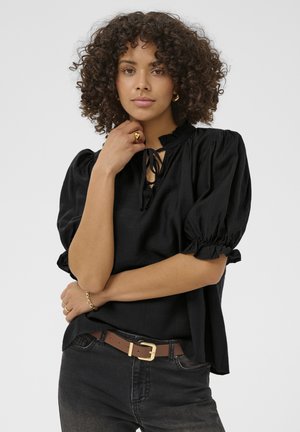 Femme aux cheveux bouclés portant un blouse noire à manches bouffantes nouée au cou, un jean foncé et une ceinture marron avec une boucle dorée sur fond uni.