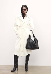 Fehér trench coat nagy hajtókákkal, övvel ellátott derékkal, és oldalzsebekkel; combközépig érő fekete lakk csizmákkal és fekete táskával párosítva.