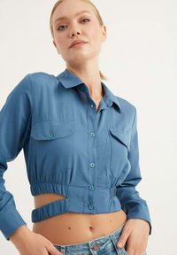 Camicetta blu corta con colletto, dotata di due tasche sul petto, vita elastica e design con tagli. Realizzata in un tessuto liscio.