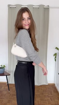 Hvid skuldertaske med kædestrop, glat tekstur. Grå langærmet top, sorte højtaljede bukser, trægulvsbaggrund, planter i nærheden.