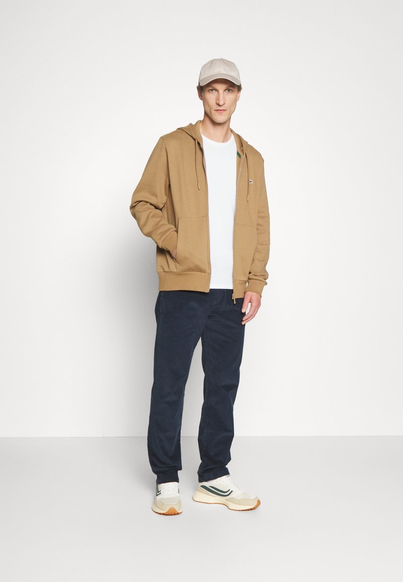 Haut à zip marron avec poches avant, t-shirt blanc en dessous, associé à un pantalon bleu foncé et des baskets multicolores, sur fond blanc.