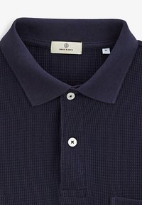 Navy blauwe poloshirt van textuurstof met een geribde kraag. Heeft een drie-knopen sluiting en witte knopen met zwarte stiksels.