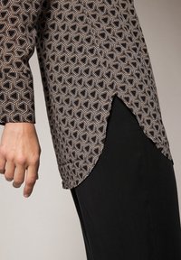 Blouse à manches longues avec motif géométrique en noir, blanc et orange. Présente un ourlet asymétrique et une texture de tissu lisse.