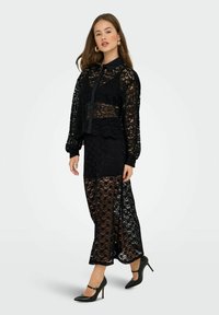 Donna che indossa una blusa nera in pizzo con maniche lunghe e bottoni, abbinata a una gonna trasparente in pizzo nera, il tutto con tacchi alti neri.