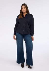 Camisa de botones de manga larga azul marino con cuello, combinada con pantalones de mezclilla oscuros de pierna ancha y botas de tobillo con tacón, mostrando una textura suave.