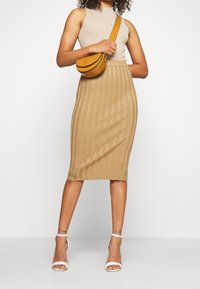 Béžový žebrovaný pletený top na ramínka v kombinaci s béžovou žebrovanou tužkovou sukní do půli stehen. Doplňky zahrnují žlutou kabelku přes rameno a bílé podpatky.