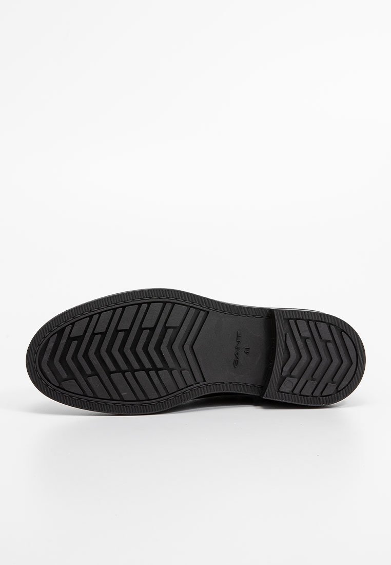 Suela de zapatilla de goma negra con un patrón de espiga texturizado, que presenta una marca y un borde elevado. Superficie lisa con contornos uniformes.