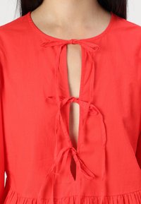 Blusa rossa con scollo rotondo e tre legature in tessuto sul davanti. Il tessuto è morbido, con una consistenza liscia e una leggera lucentezza.